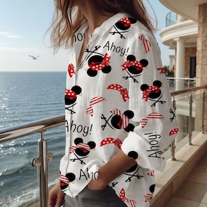 May include: White button-down shirt with a repeating pattern of black mouse-shaped pirate designs, red polka dot bows, and the word "Ahoy!" in black. The shirt has long sleeves and is worn casually.