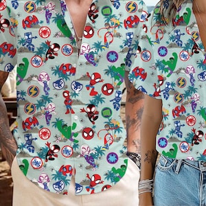 Könnte beinhalten: Zwei hellblaue Hemden mit kurzen Ärmeln, weißem Hintergrund und einem bunten Muster aus Comic-Superhelden. Das Muster enthält Rot, Blau, Grün, Gelb und Schwarz.