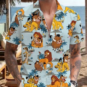 Puede incluir: Una camisa de manga corta de botones azul claro con un patrón repetido de leones, hienas y jabalíes de dibujos animados de la película El Rey León. La camisa tiene un corte clásico y un cuello.