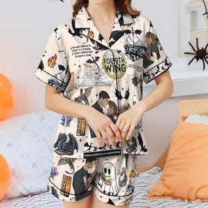 May include: Cream-colored pajama set with a short-sleeved top and shorts. The set features a repeating pattern of dragons, books, and the text "Fourth Wing." Black trim outlines the edges. The design includes quotes and illustrations, creating a fantasy theme.