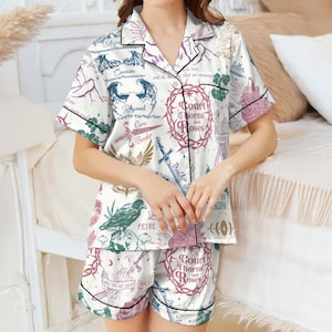 Op de afbeelding: Witte pyjamaset met een overhemd met korte mouwen en een korte broek. De set heeft een herhalend patroon van illustraties en tekst, waaronder de woorden "Court of Thorns and Roses". Zwarte biesjes omlijnen de randen van de set.