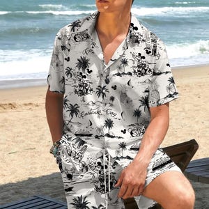 May include: White button-down shirt and matching shorts set with a black tropical print featuring palm trees, pineapples, and cartoon characters. The shirt has short sleeves and a classic collar. The shorts have a drawstring waist. The set is worn on a beach.
