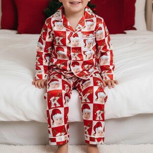 May include: A child wearing a red and white Christmas-themed pajama set. The pajamas feature a repeating pattern of baby faces wearing Santa hats. The set includes a button-down shirt and matching pants. The child is smiling and sitting on a white bed.