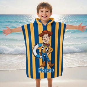 May include: A hooded beach towel featuring a cartoon image of Woody from Toy Story, with blue and yellow vertical stripes. The towel has the name "Mateo" printed on it. The child is standing on a sandy beach with the ocean in the background.