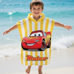 K&ouml;nnte beinhalten: Ein gelb-wei&szlig; gestreiftes Kapuzen-Badetuch mit einem roten Cartoon-Rennwagen mit der Nummer 95. Auf dem Handtuch steht der Name "Mateo". Das Kind steht an einem Sandstrand mit dem Meer im Hintergrund.