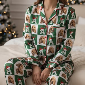 May include: Green and white pajama set with a repeating pattern of brown dogs wearing Santa hats and the letter "A". The pajamas have red trim and are made of a silky material. The model is sitting on a bed.