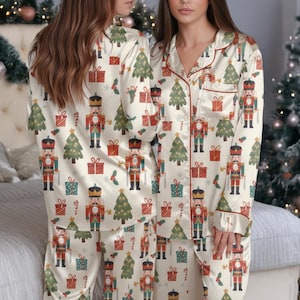 May include: Cream-colored pajama set with a festive Christmas pattern. The long-sleeved top and pants feature a repeating design of nutcrackers, Christmas trees, and wrapped gifts. Red trim accents the collar and cuffs, creating a cozy holiday look.