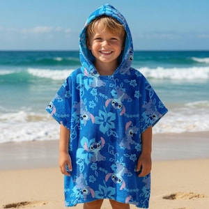 Puede incluir: Un poncho de playa azul con capucha con un patr&oacute;n repetitivo de un personaje de dibujos animados y dise&ntilde;os florales. El poncho lo lleva un ni&ntilde;o en una playa de arena con el oc&eacute;ano de fondo.