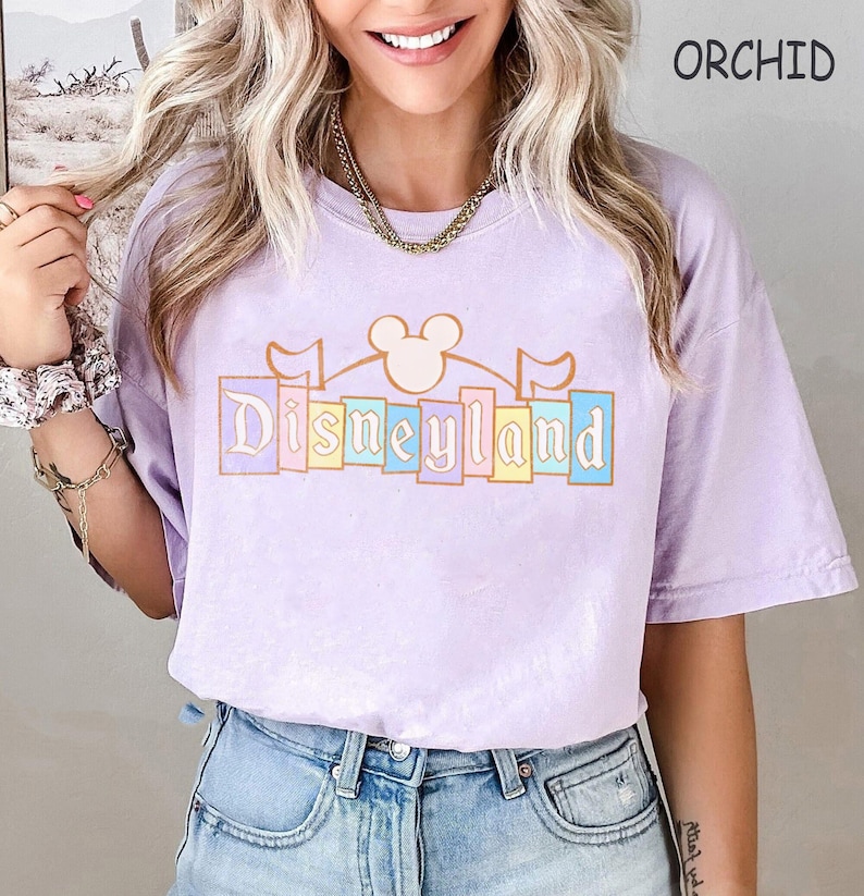 May include: A lilac t-shirt featuring the word "Disneyland" in a pastel design, with a Mickey Mouse head and flags. The shirt has a relaxed fit and is made of a soft material. The word "ORCHID" is printed on the top right.