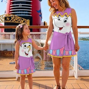 Puede incluir: Dos vestidos sin mangas con un gráfico del personaje Sra. Potts de La Bella y la Bestia. Los vestidos tienen un corpiño blanco con la cara del personaje, una falda morada con lazo y un dobladillo festoneado con detalles en rosa y azul.