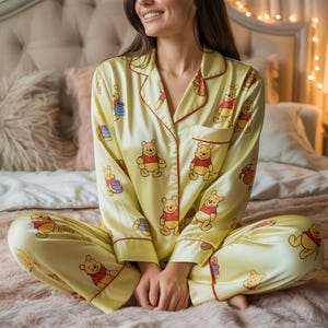 Puede incluir: Conjunto de pijama de satén amarillo con un estampado repetido de un oso de dibujos animados con una camisa roja. El pijama tiene ribetes rojos y una camisa abotonada con un bolsillo en el pecho. El pantalón y la camisa son visibles.