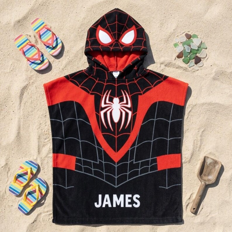 May include: A black and red hooded poncho towel featuring a spiderweb pattern, a white spider emblem on the chest, and white eyes on the hood. The name JAMES is printed in white capital letters at the bottom of the towel, which is laid flat.
