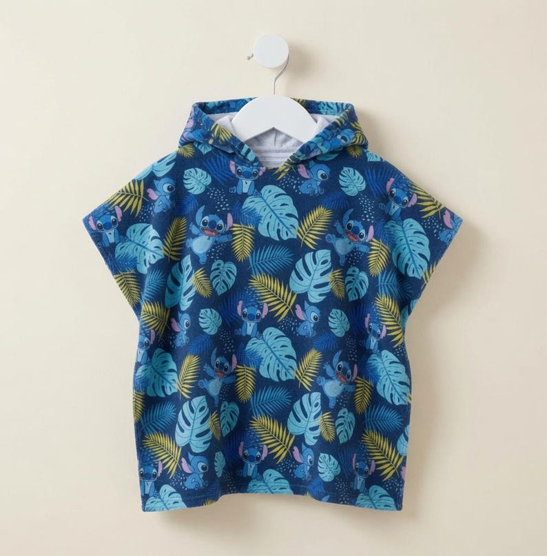 Puede incluir: Una toalla tipo poncho con capucha que presenta un fondo azul oscuro con un estampado integral del personaje Stitch en varias poses, intercalado con hojas de monstera azul claro y frondas de palma amarillas, colgada en una percha blanca.