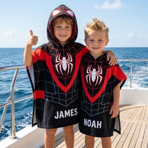 May include: Two black and red hooded poncho towels featuring a white spider web graphic and a central white spider emblem. The left towel is personalized with the name JAMES in white text, and the right towel is personalized with the name NOAH.