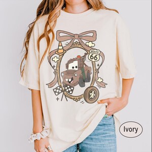May include: Ivory-colored t-shirt featuring a cartoon graphic of Tow Mater from the movie Cars, framed by a brown ribbon and Route 66 sign. The design includes a traffic cone, checkered flags, and a tire. The shirt is a relaxed fit.