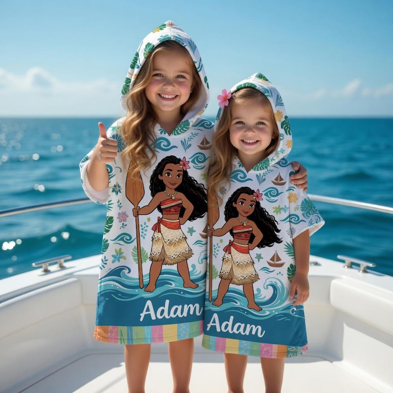 K&ouml;nnte beinhalten: Zwei Kapuzen-Badet&uuml;cher mit einem Cartoon-Charakter-Design. Die Handt&uuml;cher sind wei&szlig; mit einem blauen und t&uuml;rkisfarbenen Wellenmuster, und die Figur h&auml;lt ein Holzpaddel. Der Name "Adam" ist unten aufgedruckt.