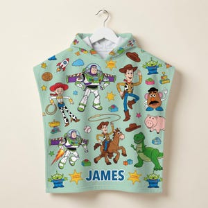 May include: A light green hooded towel featuring a repeating pattern of Toy Story characters including Buzz Lightyear, Woody, Jessie, Rex, and Mr. Potato Head. The name JAMES is printed in blue capital letters near the bottom center of the towel.