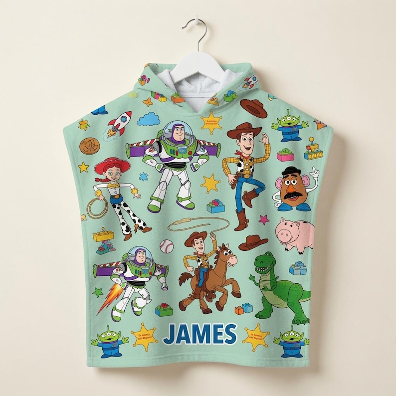 May include: A light green hooded towel featuring a repeating pattern of Toy Story characters including Buzz Lightyear, Woody, Jessie, Rex, and Mr. Potato Head. The name JAMES is printed in blue capital letters near the bottom center of the towel.