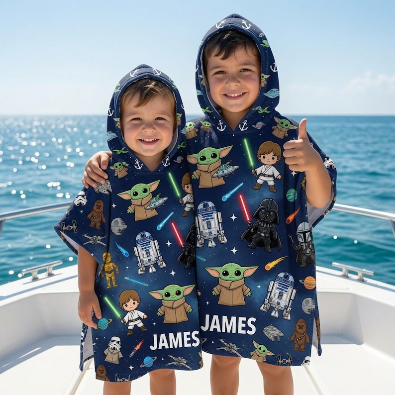 May include: Two navy blue hooded poncho towels featuring a repeating Star Wars character print including Grogu, Luke Skywalker, Darth Vader, and R2-D2. The fabric is decorated with white anchors and the name JAMES printed in white capital letters.