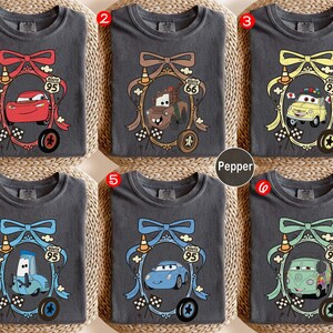 May include: Six charcoal gray t-shirts with different cartoon car designs inside decorative frames. Each frame has a bow, Route 95 sign, and a tire. The designs include a red car, a brown truck, a yellow car, a blue car, and a green van.
