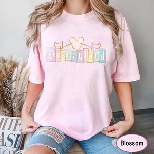 May include: A pale pink t-shirt featuring the word "Disneyland" in pastel shades. The design incorporates a Mickey Mouse silhouette and flags. The shirt has a relaxed fit.