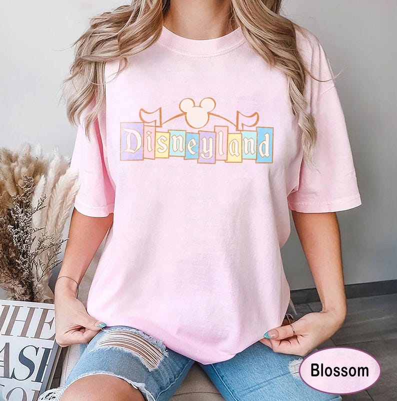 May include: A pale pink t-shirt featuring the word "Disneyland" in pastel shades. The design incorporates a Mickey Mouse silhouette and flags. The shirt has a relaxed fit.