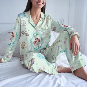 May include: Light green pajama set featuring a repeating pattern of Ariel from The Little Mermaid, seashells, and other aquatic elements. The long-sleeved top has a notched collar and button closure. The pants have an elastic waistband and a relaxed fit.