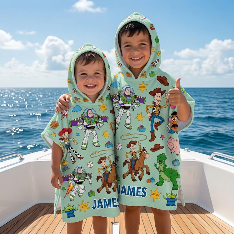 May include: Two light green hooded poncho towels featuring a repeating pattern of Toy Story characters including Buzz Lightyear, Woody, Jessie, Rex, and Mr. Potato Head, with the name JAMES printed in blue on the front of each garment.