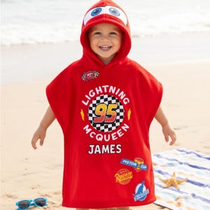 May include: A red Lightning McQueen hooded beach towel with a hood featuring car eyes. The towel has the text "LIGHTNING MCQUEEN 95 JAMES" and various car-themed patches. The child is on a sandy beach.