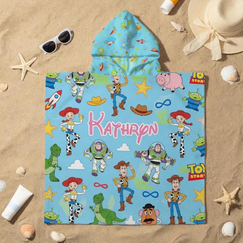 May include: A light blue hooded beach towel featuring characters from the Toy Story movie, including Woody, Buzz Lightyear, and Jessie. The name "Kathryn" is printed in pink script. The towel is decorated with stars, hats, and other themed graphics.