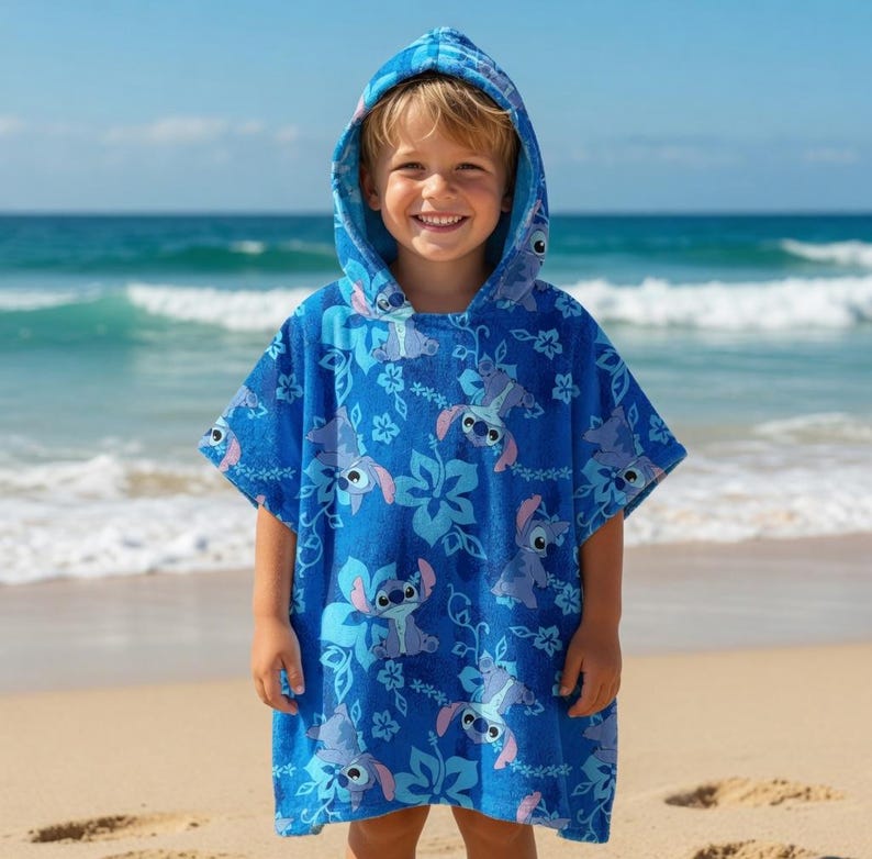 Puede incluir: Un poncho de playa azul con capucha con un patr&oacute;n repetitivo de un personaje de dibujos animados y dise&ntilde;os florales. El poncho lo lleva un ni&ntilde;o en una playa de arena con el oc&eacute;ano de fondo.