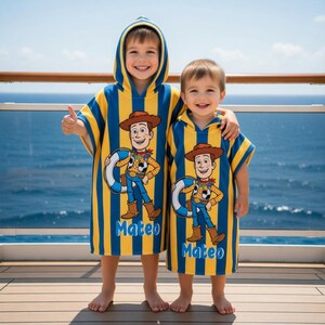 May include: Two children wearing matching hooded beach towels. The towels are blue and yellow striped with a Woody cartoon character design and the name "Mateo" printed on them. The children are smiling and standing on a wooden deck with a blue ocean in the background.
