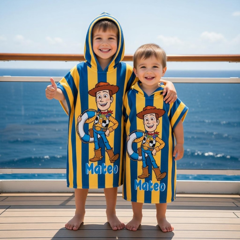 May include: Two children wearing matching hooded beach towels. The towels are blue and yellow striped with a Woody cartoon character design and the name "Mateo" printed on them. The children are smiling and standing on a wooden deck with a blue ocean in the background.