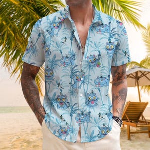 May include: Light blue short-sleeve button-down shirt featuring a repeating pattern of cartoon cats and palm trees. The cats are in various poses and expressions. The shirt is worn open, revealing a silver necklace.