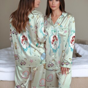 May include: Light green satin pajama set featuring a repeating pattern of Ariel from The Little Mermaid, seashells, and other aquatic-themed illustrations. The long-sleeved top and pants set has a button-down shirt with green trim.