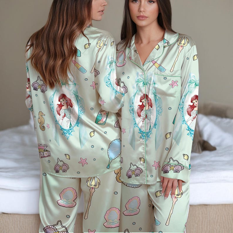 May include: Light green satin pajama set featuring a repeating pattern of Ariel from The Little Mermaid, seashells, and other aquatic-themed illustrations. The long-sleeved top and pants set has a button-down shirt with green trim.
