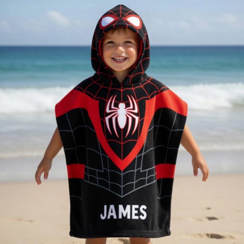 May include: A black hooded poncho towel featuring a red and black spiderweb pattern, a large white spider emblem on the chest, and white eye details on the hood. The name JAMES is printed in white block letters at the bottom of the garment.