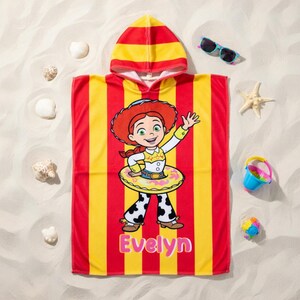 May include: A hooded beach towel featuring a cartoon character in a yellow and red striped design. The towel has the name "Evelyn" printed in pink. Additional beach accessories include sunglasses, a bucket, and seashells.