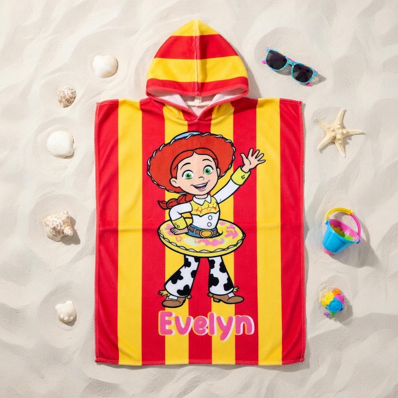May include: A hooded beach towel featuring a cartoon character in a yellow and red striped design. The towel has the name "Evelyn" printed in pink. Additional beach accessories include sunglasses, a bucket, and seashells.