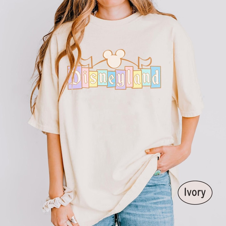 May include: Ivory-coloured t-shirt featuring the word "Disneyland" in pastel shades. The design incorporates a Mickey Mouse head and flags. The shirt has a relaxed fit and short sleeves.