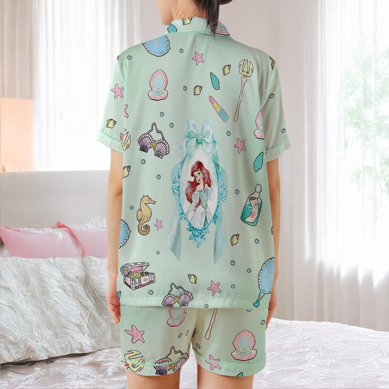 May include: Light green pajama set featuring a print of Ariel from The Little Mermaid, seashells, and other aquatic-themed illustrations. The set includes a short-sleeved button-up shirt and matching shorts. The shirt has a large framed image of Ariel on the back.