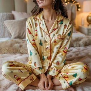 May include: Yellow satin pajama set featuring a Winnie the Pooh Christmas print. The long-sleeved top and pants have red trim and are decorated with cartoon characters, Christmas trees, and candy canes. The pajama set is made of a soft, silky material.
