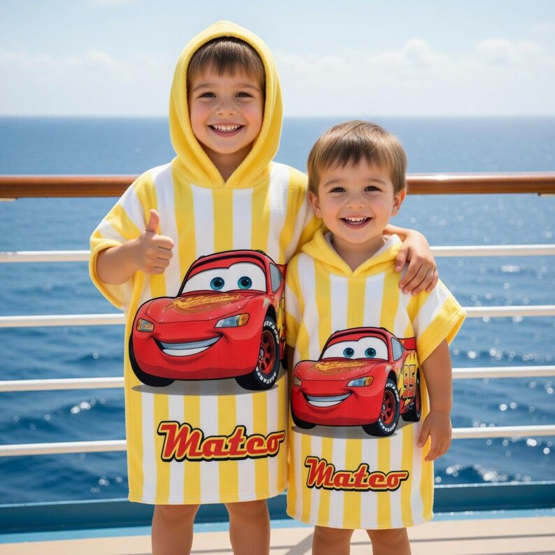 K&ouml;nnte beinhalten: Zwei Kinder tragen gelb-wei&szlig; gestreifte Kapuzen-Badet&uuml;cher mit einem roten Lightning McQueen-Auto-Motiv. Auf den Handt&uuml;chern steht das Wort "Mater". Die Kinder l&auml;cheln vor dem Meer.