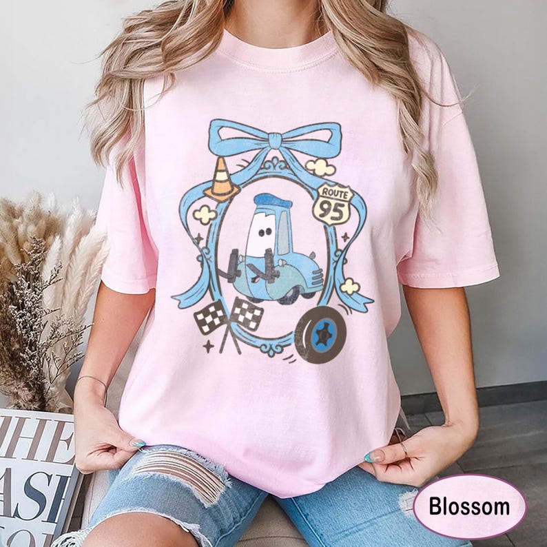 May include: A light pink t-shirt featuring a cartoon car design within a blue ribbon frame. The design includes a Route 95 sign, checkered flags, and a tire. The shirt is a comfortable, casual style.