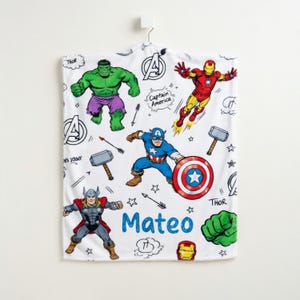 May include: White superhero-themed blanket featuring illustrations of Hulk, Iron Man, Captain America, and Thor. The blanket includes the name "Mateo" in blue, along with comic book style elements.