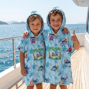 May include: Two light blue hooded beach towels featuring a repeating pattern of the character Stitch from Lilo and Stitch, palm trees, and tropical holiday scenes. The towels are designed as poncho-style garments with hoods and open sides.