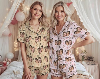 May include: Two women wearing matching pajama sets. One set is light yellow with black trim, and the other is light pink with black trim. Both sets feature a pattern of faces and hearts. The pajamas have short sleeves and shorts.