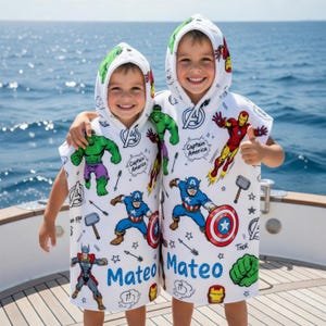 May include: Two children wearing white hooded beach towels featuring Marvel comic book characters. The towels have images of Captain America, Iron Man, Thor, and the Hulk. The name "Mateo" is printed in blue on each towel.