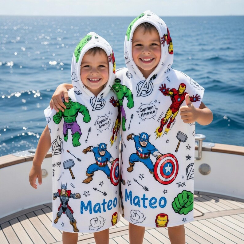May include: Two children wearing white hooded beach towels featuring Marvel comic book characters. The towels have images of Captain America, Iron Man, Thor, and the Hulk. The name "Mateo" is printed in blue on each towel.
