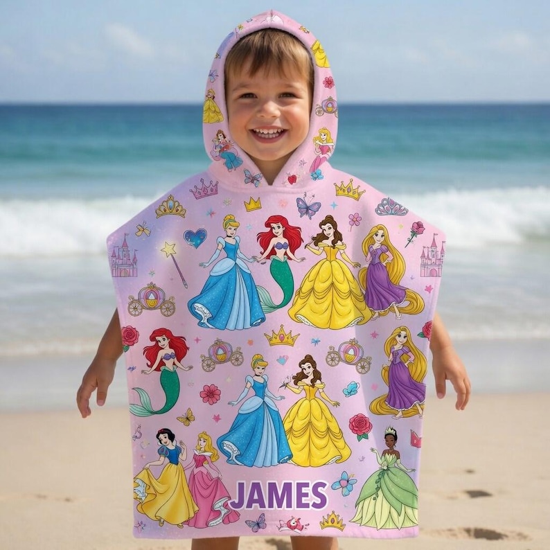 Puede incluir: Una toalla con capucha de color rosa claro que presenta un patr&oacute;n repetitivo de varias princesas Disney, incluyendo a Ariel, Cenicienta, Bella y Rapunzel, junto con coronas, castillos y carrozas. El nombre JAMES est&aacute; impreso en morado en la parte inferior central.